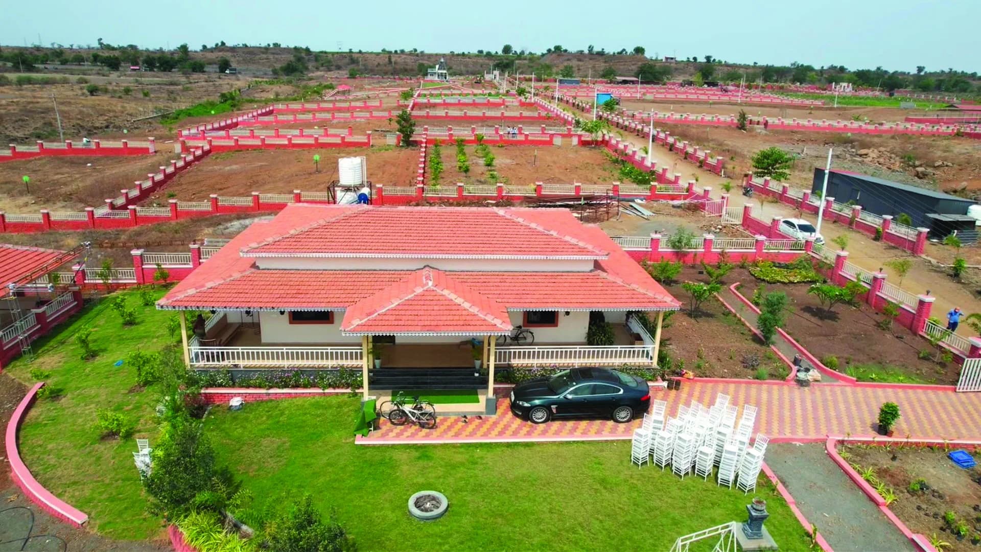 Aerial photo of Isha Farms: a completed farmhouse with a red-tile roof amid demarcated plots with pink boundary walls